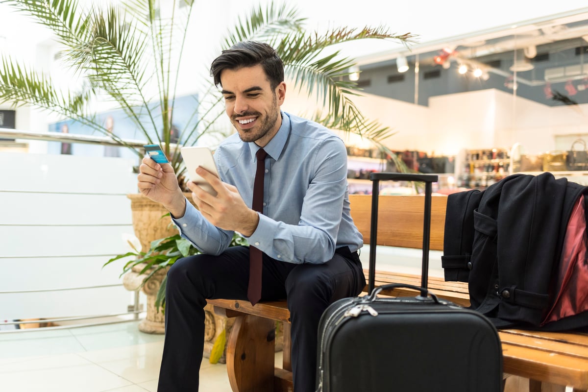 A person holding a payment card and looking at a mobile phone while sitting with luggage at an airport.