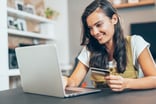 A person holding a payment card and smiling while sitting in-front of a laptop computer.