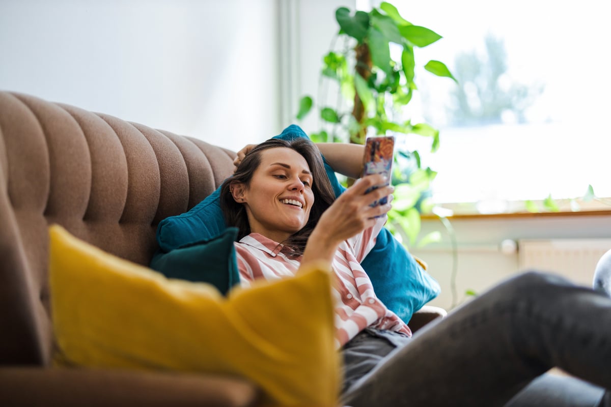 A person smiling while laying on a couch and looking at a mobile phone.