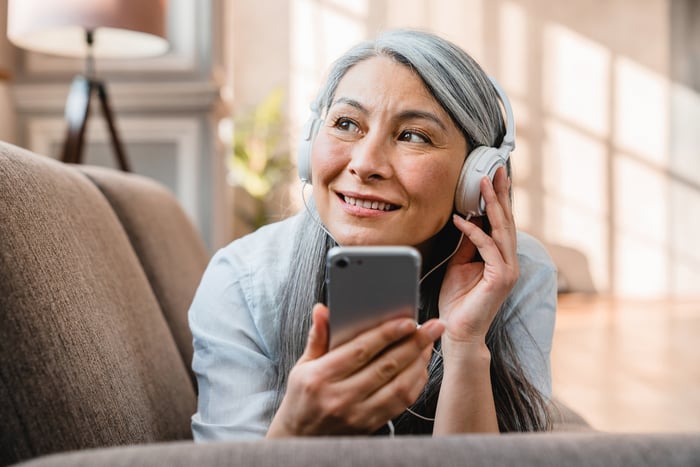 A person smiling while lying on a couch, wearing headphones and holding a mobile phone.