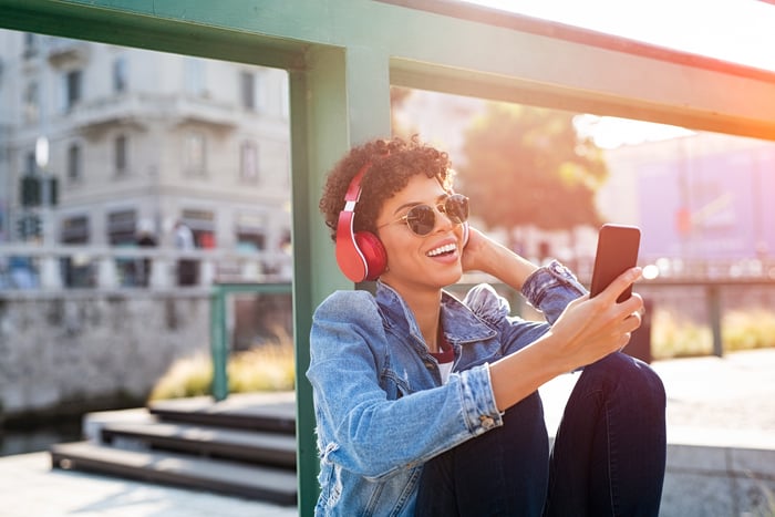 A person sitting outdoors looks at a mobile phone while smiling.