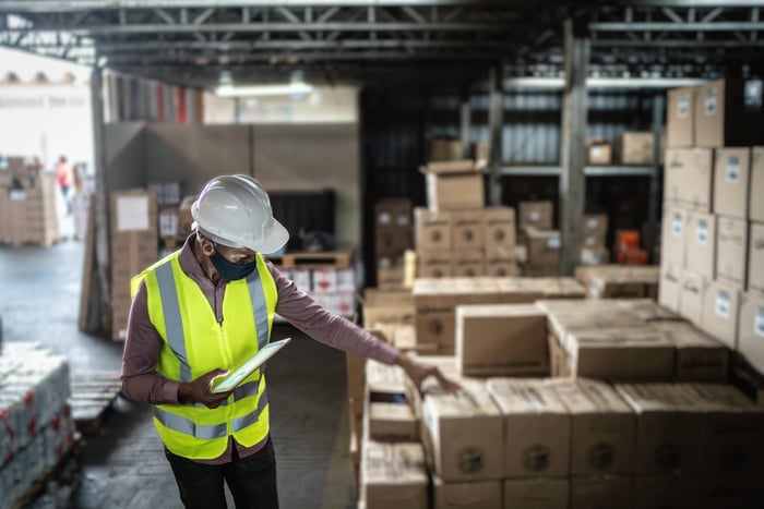 A person holding a clipboard and wearing personal protective equipment looking at a stack of packages for shipment.