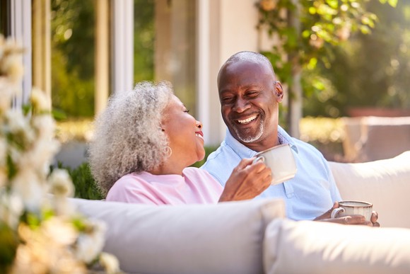 Two people sitting on a couch outdoors drinking from mugs and smiling at each other.