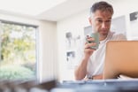 A person holding a mug and looking at a laptop computer screen in a serious manner.