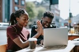 Two people smile and high-five while sitting at an outdoor table in-front of a laptop computer.