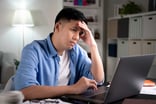 A person puts their hand on their forehead while looking at a computer in a concerned manner.