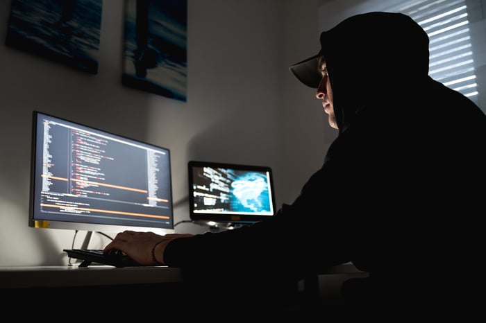 A computer hacker in a black hoodie sweatshirt typing on a keyboard in a dimly-lit room.