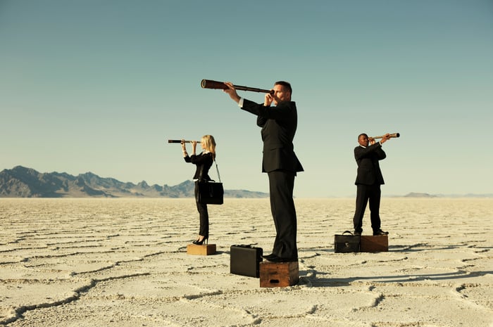 Three people standing on boxes in a desert looking through telescopes.