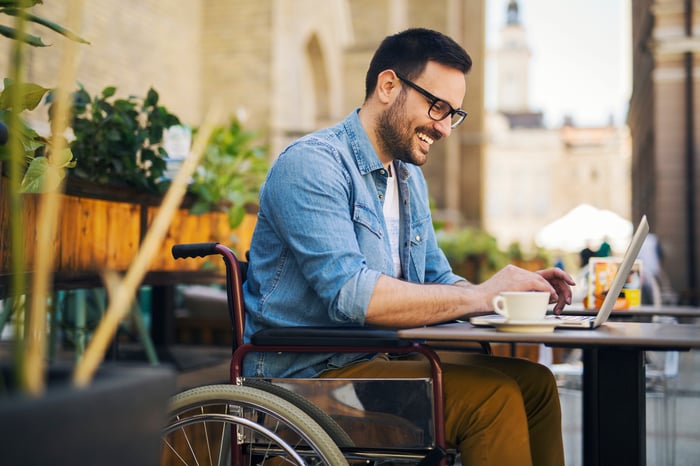 A person smiling while sitting at a table outside and typing on a laptop computer.