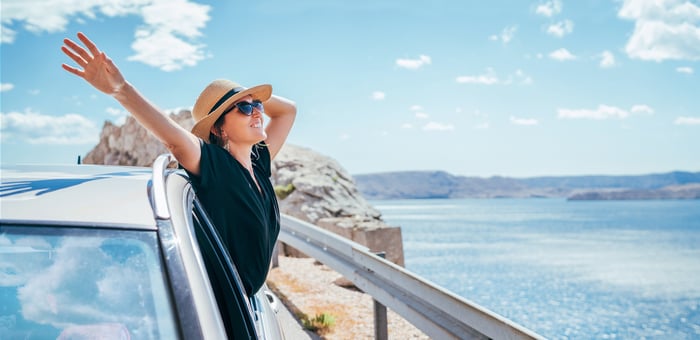 A person smiling while leaning out of a car window by a body of water.