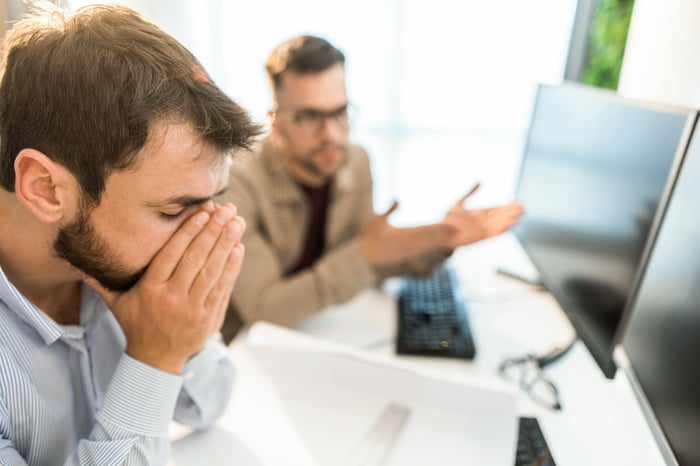 A person sits with face in hands while a colleague gestures with their hands.