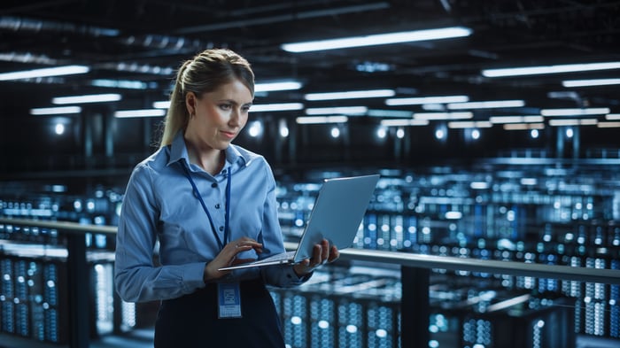 A technician checks her laptop inside a large data center room filled with computer servers.