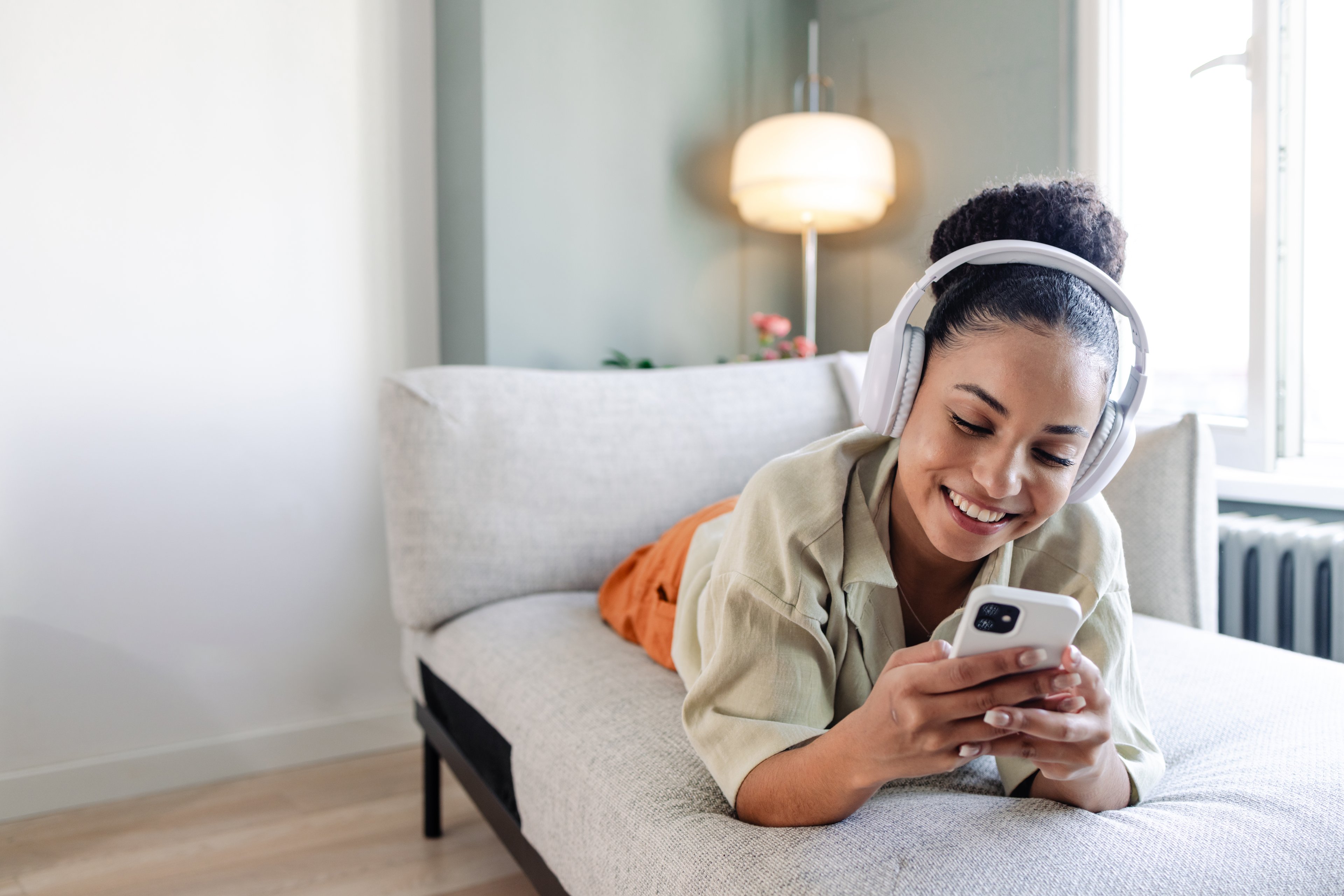 A smiling person laying on the couch with headphones on, and smartphone in hand