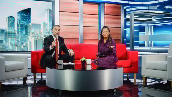 Two TV broadcasters sit in a studio set.