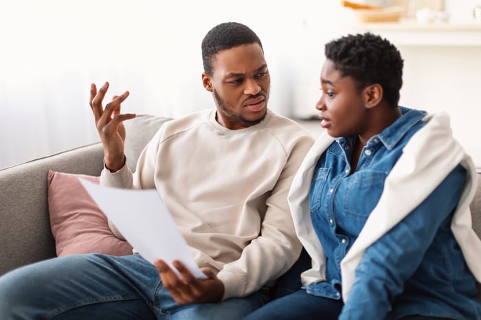 Two people sitting on a couch reacting to information on a sheet of paper in a confused manner.