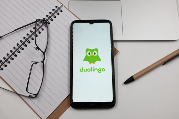 The Duolingo logo appears on a smartphone sitting on a desk alongside glasses, a notebook, a pen, and a closed laptop.