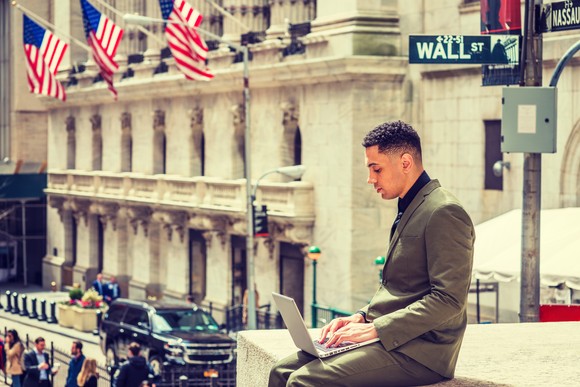 A person sitting in front of the Wall Street sign and working on a laptop.