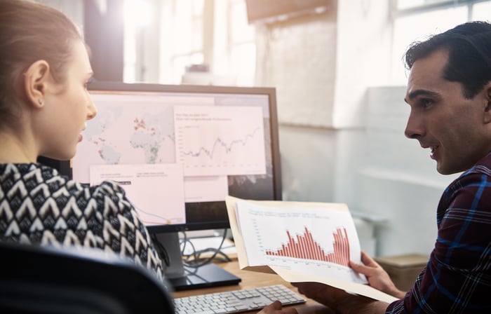 A pair of investors sit in front of a computer screen while holding several print outs of stock charts and discussing them with each other.