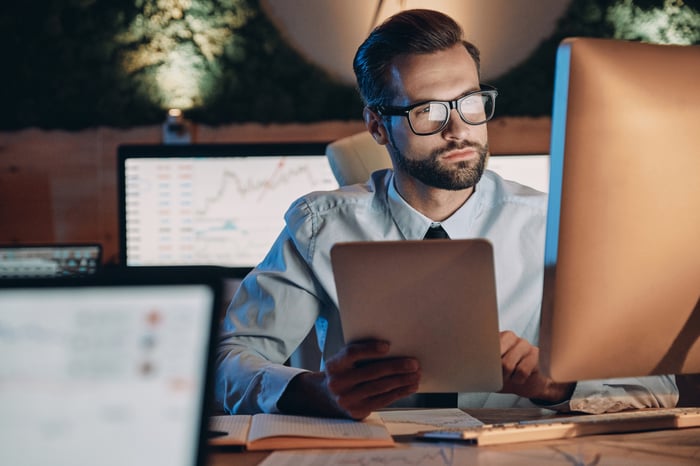 Very serious person sitting at a desk working on a computer with stock charts visible on the computer screens behind him. 
