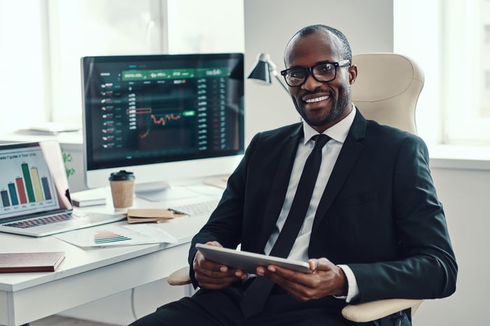 Businessperson smiles while holding a tablet. A computer screen showing a stock chart is in the background.