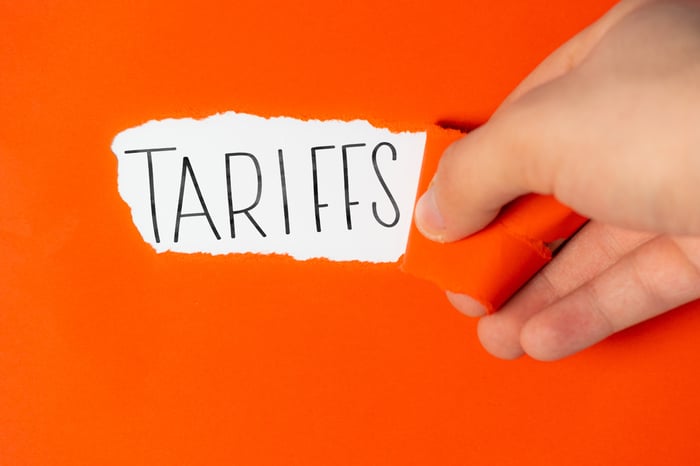 A hand tearing orange paper away to reveal the word "Tariffs."