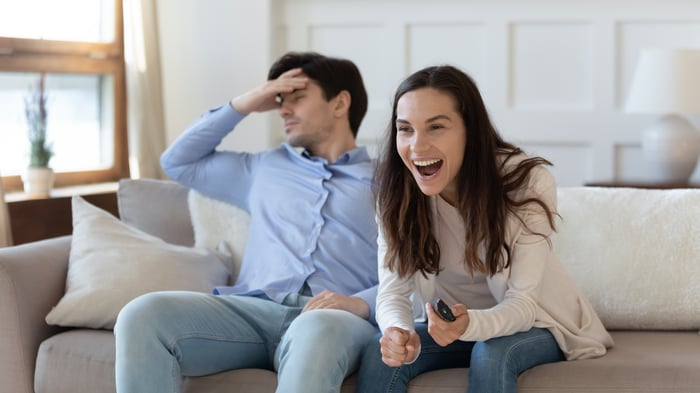 Two people on the TV couch, displaying opposite moods. One is laughing at the screen while the other looks away, frowning.