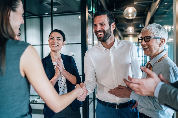 Smiling businesspeople shaking hands while others applaud.