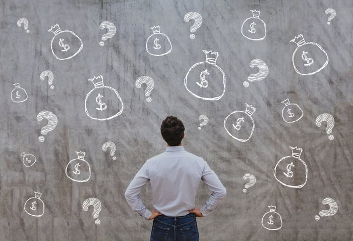 Investor stares at screen with dollar bags and question marks.