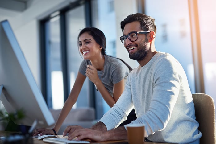 Two people smile while they look at something on a computer in an office.
