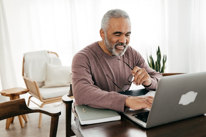 A person smiles while holding their glasses and sitting at a table and typing on a laptop computer.