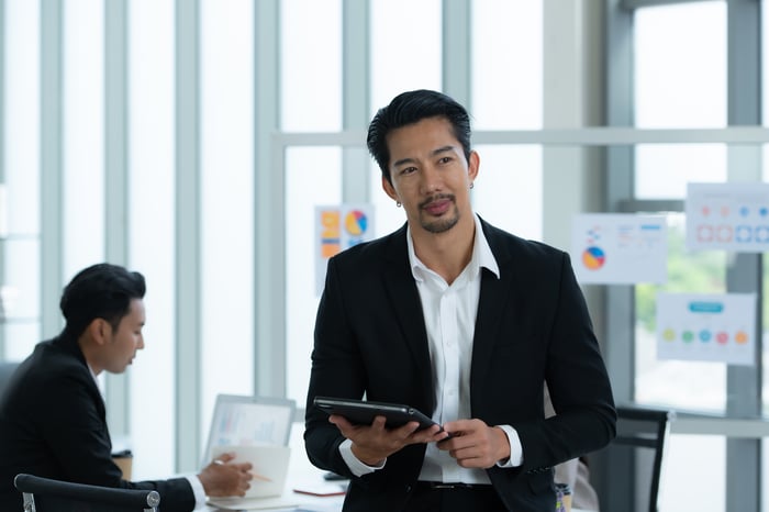 A person holding a tablet in an office setting and looking thoughtful.