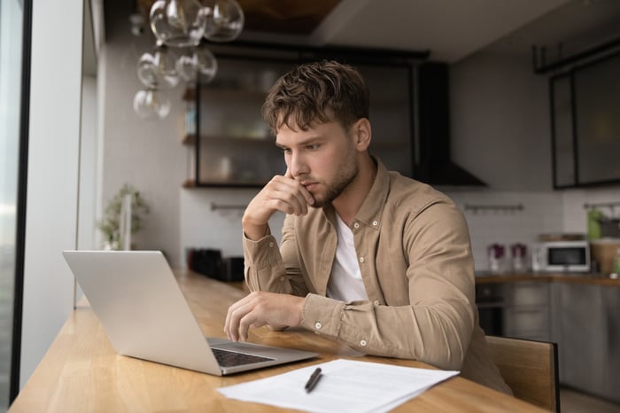 A person looking thoughtfully at a computer while sitting in their kitchen.