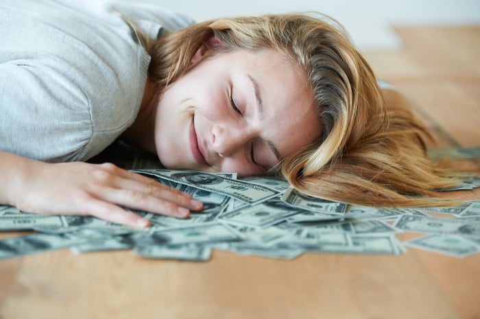 A smiling person with their head lying on lots of cash strewn across a table.