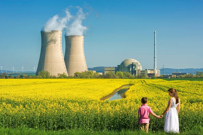 Two people, holding hands, look across a field of yellow flowers toward a nuclear power plant at the horizon. 