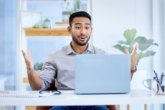 A person with a surprised expression and outstretched hands looking at a laptop.