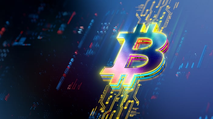 A rainbow-colored Bitcoin logo rests on a circuit board and tiling stock chart patterns.