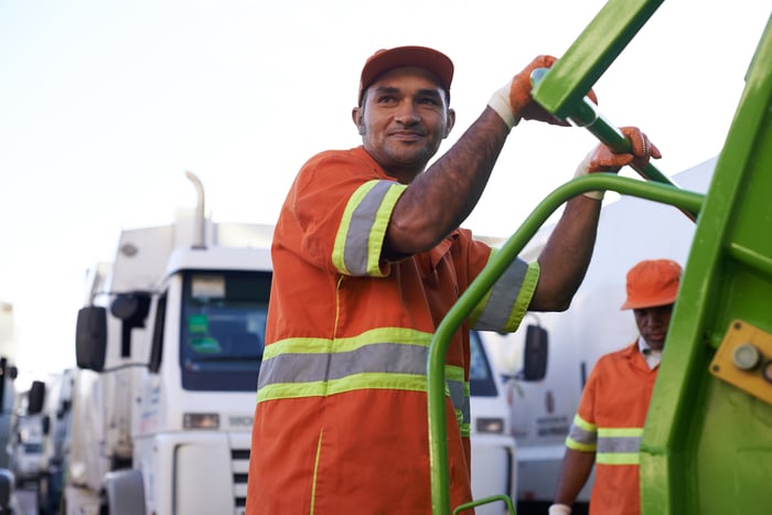 A person smiling while holding onto a sanitation truck for waste pickup.