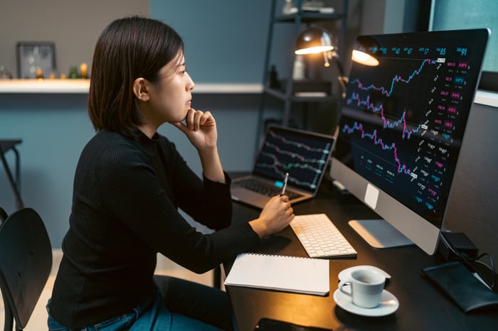 A person looking at stock charts on a desktop and a laptop.