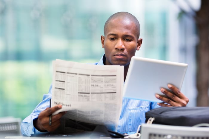 Middle-aged man sitting down while reading a business newspaper and using a tablet. 