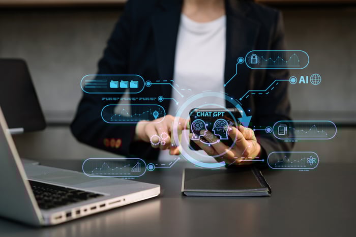 Someone using a cellphone while seated before a laptop, with a ChatGPT graphic superimposed.