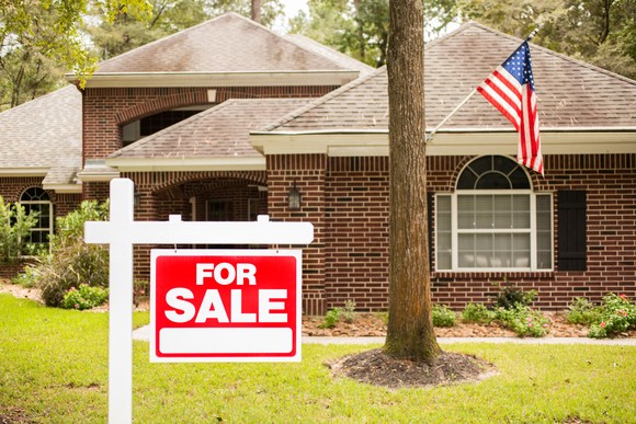 A for sale sign in front of a house with an American flag protruding from a tree on the front lawn.