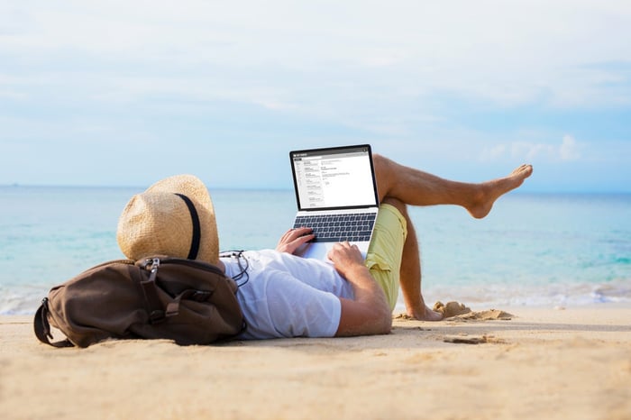 Man lying on a beach with the ocean on the background, working on a laptop. 