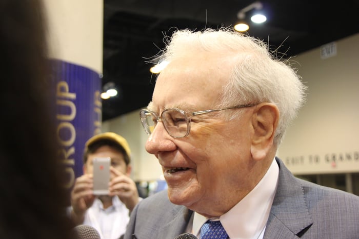 Warren Buffett smiling with a person taking a photo in the background.