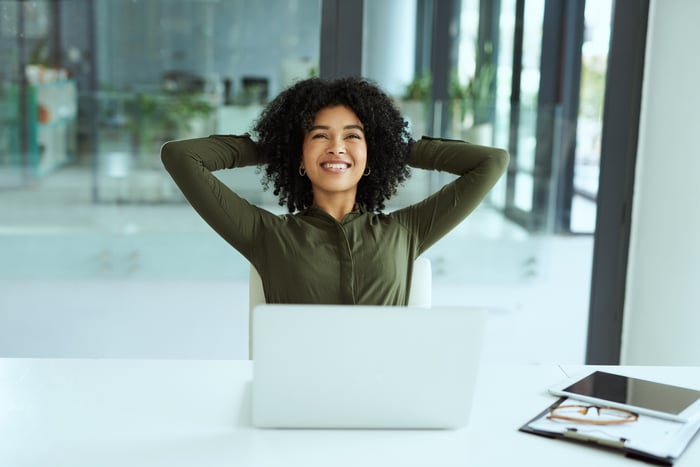 A smiling person sitting in front of a laptop while holding hands behind head.
