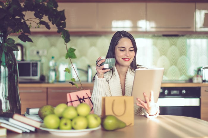 A person in a kitchen looks at a tablet while holding a mug of tea.