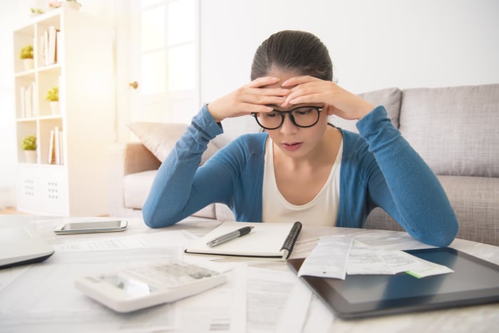 An investor touches her head in frustration while sitting at a table covered with notebooks and papers.