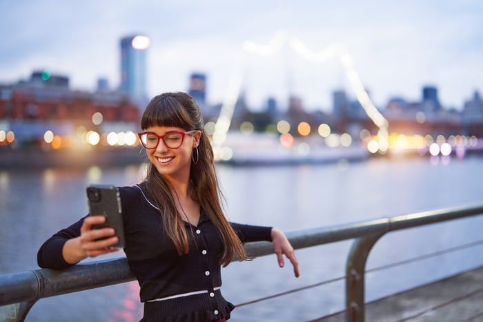 A person smiles while looking at their cell phone and leaning on a railing in an urban setting next to a river.