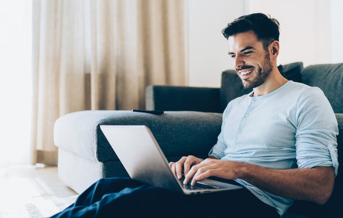 A person smiles while sitting against a couch and typing on a laptop computer.