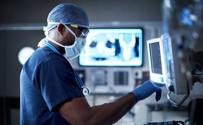 Someone in scrubs and mask operating medical monitoring equipment.