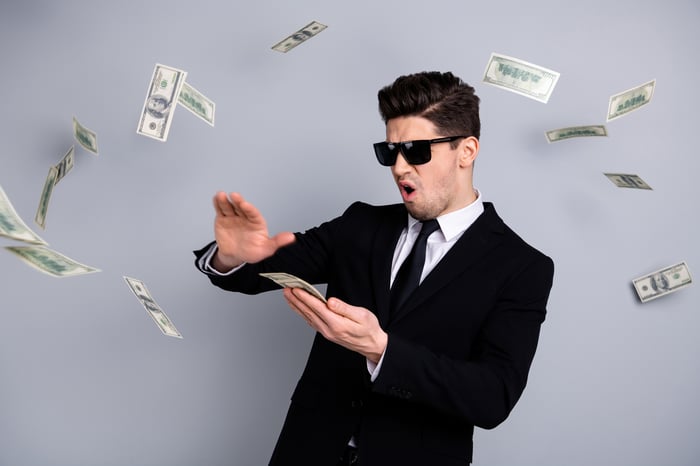 A person in a black suit and shades peeling off $100 bills.
